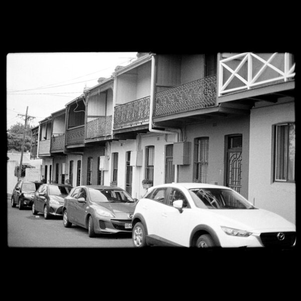 A series of parked cars on a city street lined with townhouse buildings in black and white.